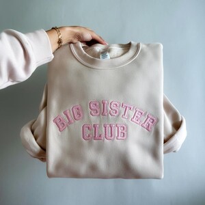 K&ouml;nnte beinhalten: Ein beiger Pullover mit einem rosa gestickten Bogen, auf dem "BIG SISTER CLUB" steht.