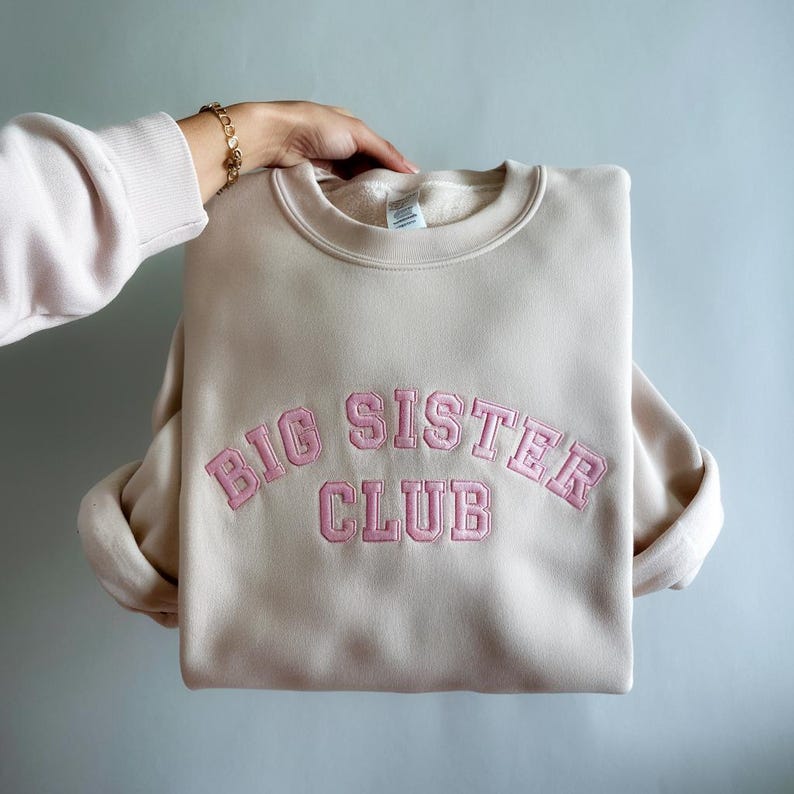K&ouml;nnte beinhalten: Ein beiger Pullover mit einem rosa gestickten Bogen, auf dem "BIG SISTER CLUB" steht.
