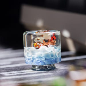 Puede incluir: Un pisapapeles decorativo de resina transparente con un barco en miniatura navegando sobre formaciones de olas azules y blancas. El barco tiene una vela blanca con un personaje de dibujos animados y una vela a rayas rojas y naranjas.