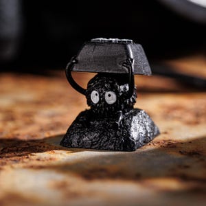 Pode incluir: Um keycap preto e branco com um design de criatura peluda. A criatura tem dois olhos grandes e está segurando um keycap preto acima de sua cabeça.