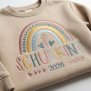 May include: A beige crewneck sweatshirt featuring an embroidered design of a multi-colored rainbow with leaf accents, two hearts, and an ice cream cone. Below the rainbow, the text SCHULKIND 2026 is embroidered in multi-colored lettering.
