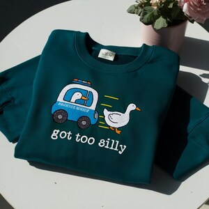 May include: Teal crew neck sweatshirt with a cartoon duck in a police car chasing another duck. The text "got too silly" is printed below the graphic. The sweatshirt is made of a soft material.