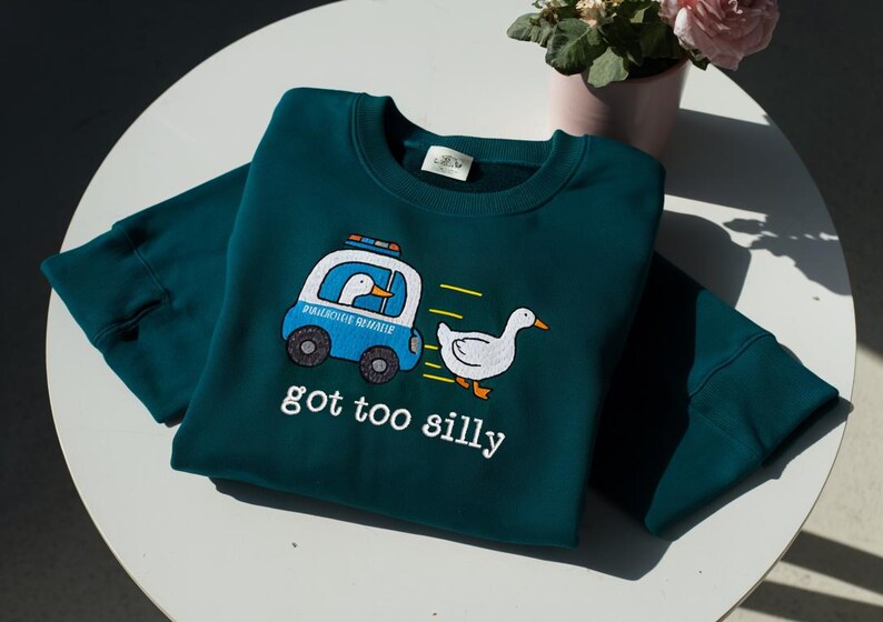 May include: Teal crew neck sweatshirt with a cartoon duck in a police car chasing another duck. The text "got too silly" is printed below the graphic. The sweatshirt is made of a soft material.