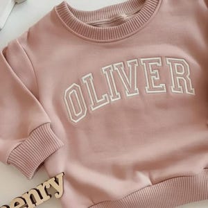 Puede incluir: Sudadera rosa claro con el nombre "OLIVER" bordado en letras blancas en el pecho. La sudadera tiene pu&ntilde;os y dobladillo de canal&eacute;. Las mangas son largas y el cuello es redondo.