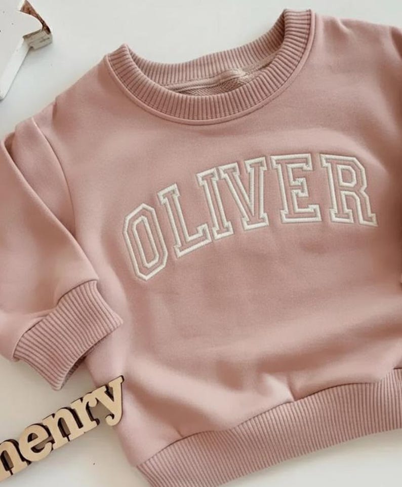 Puede incluir: Sudadera rosa claro con el nombre "OLIVER" bordado en letras blancas en el pecho. La sudadera tiene pu&ntilde;os y dobladillo de canal&eacute;. Las mangas son largas y el cuello es redondo.