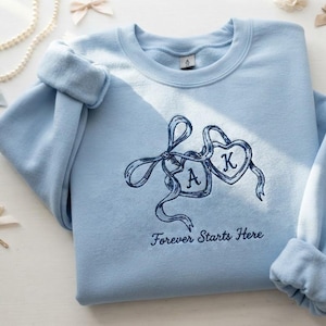 Op de afbeelding: Een lichtblauw sweatshirt met ronde hals en donkerblauw borduurwerk van twee met elkaar verbonden hartvormige medaillons vastgebonden met een lint. De letters A en K staan in het midden van de harten en de tekst Forever Starts Here staat eronder.