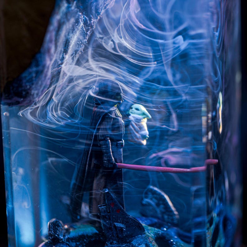 Puede incluir: Una figura oscura, posiblemente Darth Vader, se encuentra en un bloque de resina azul y blanco con patrones de humo en espiral. Una figura m&aacute;s peque&ntilde;a, posiblemente Yoda, es visible detr&aacute;s de la m&aacute;s grande.