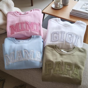 Puede incluir: Cuatro sudaderas de cuello redondo en colores pastel. Cada una tiene bordada una palabra en estilo universitario: "MIMI" en rosa, "GIGI" en blanco, "MAMA" en azul claro y "PAPA" en verde oliva. Un estilo casual y cómodo.