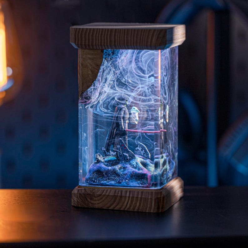 Puede incluir: Una base de l&aacute;mpara de madera con un cilindro de vidrio transparente que contiene una escena en miniatura de dos figuras de pie sobre una superficie azul y morada. El cilindro de vidrio est&aacute; lleno de remolinos blancos y una luz roja brilla desde las figuras.