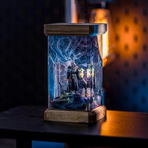 Puede incluir: Una base de l&aacute;mpara de madera con un cilindro de vidrio transparente que contiene una figura oscura y una figura m&aacute;s peque&ntilde;a. El cilindro de vidrio est&aacute; lleno de resina azul y morada con remolinos blancos. La base de la l&aacute;mpara est&aacute; iluminada desde adentro, proyectando un brillo c&aacute;lido.