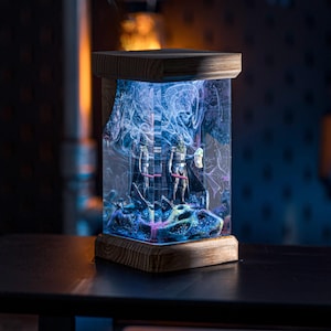 Puede incluir: Una base de l&aacute;mpara de madera con un cilindro de vidrio transparente que contiene resina azul y morada con un dise&ntilde;o gal&aacute;ctico. Dos peque&ntilde;as figuras son visibles dentro de la resina.