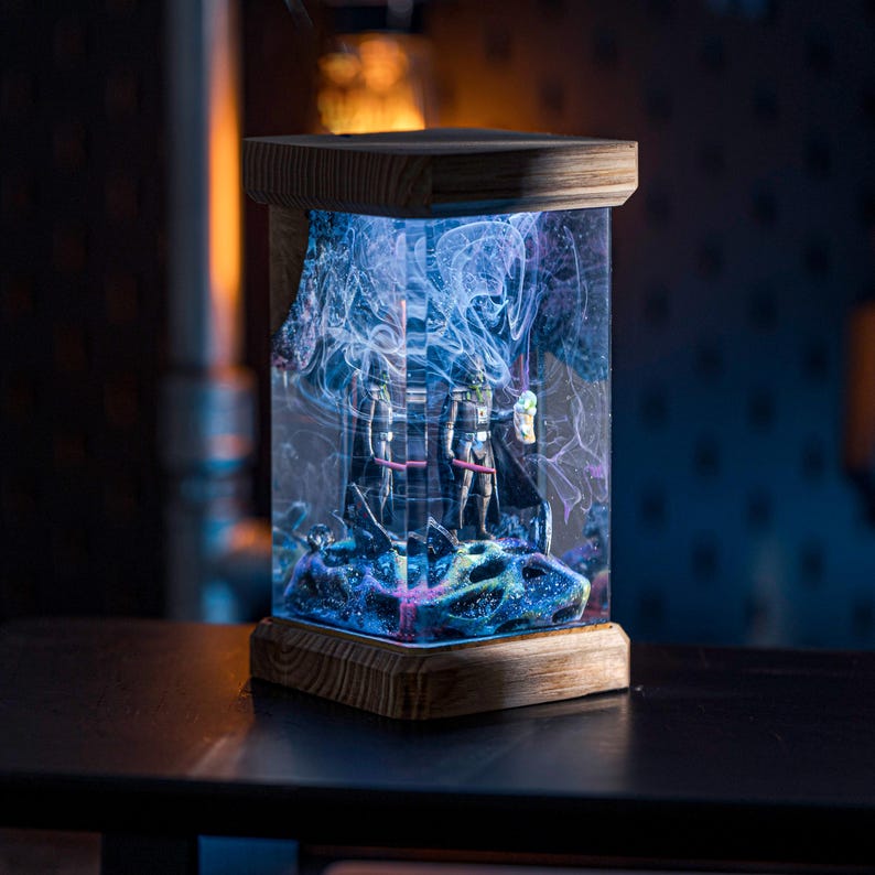 Puede incluir: Una base de l&aacute;mpara de madera con un cilindro de vidrio transparente que contiene resina azul y morada con un dise&ntilde;o gal&aacute;ctico. Dos peque&ntilde;as figuras son visibles dentro de la resina.