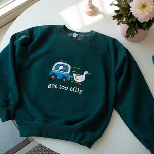 May include: A dark teal sweatshirt with a whimsical design featuring a blue vehicle with a duck inside, a running white duck, and the text "got too silly". The sweatshirt is on a white table with flowers.