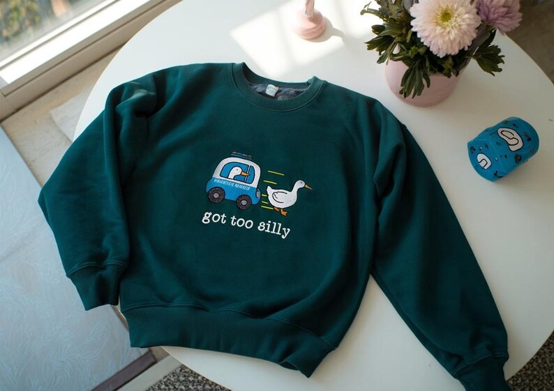May include: A dark teal sweatshirt with a whimsical design featuring a blue vehicle with a duck inside, a running white duck, and the text "got too silly". The sweatshirt is on a white table with flowers.