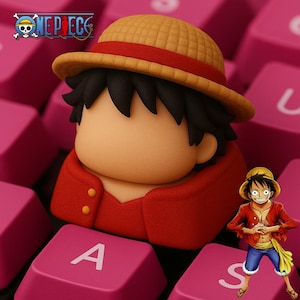 以下が含まれることがあります： ワンピースのアニメのキャラクターの赤と茶色のフィギュア。キャラクターは麦わら帽子と赤いシャツを着ています。フィギュアは、AとSの文字が見えるピンクのキーボードの上に座っています。