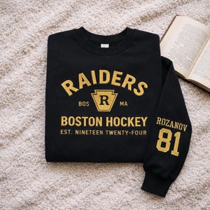 Könnte beinhalten: Schwarzes Sweatshirt mit goldfarbenem Schriftzug "RAIDERS" über "BOSTON HOCKEY" und "EST. NINETEEN TWENTY-FOUR". Der Ärmel hat "ROZANOV 81" in Gold. Das Sweatshirt liegt gefaltet auf einer strukturierten Oberfläche.