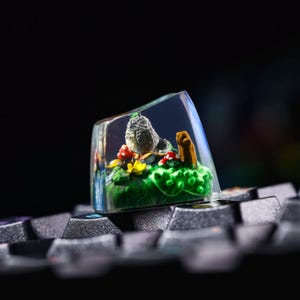 May include: A clear resin keycap with a miniature scene inside. The scene features a small gray bird, green foliage, and red and yellow mushrooms.