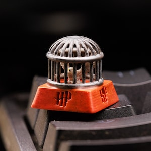 May include: A small, orange keycap with a silver cage on top. The cage contains a white owl figurine. The keycap has the letters "HP" printed on it.