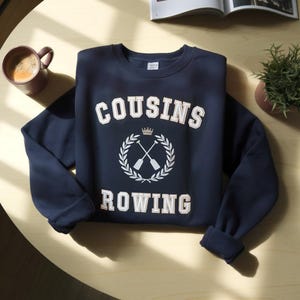 Puede incluir: Una sudadera azul marino con las palabras "COUSINS ROWING" en letras blancas. El diseño incluye una corona y remos cruzados dentro de una corona de laurel. También se ven una taza de café y una pequeña planta.