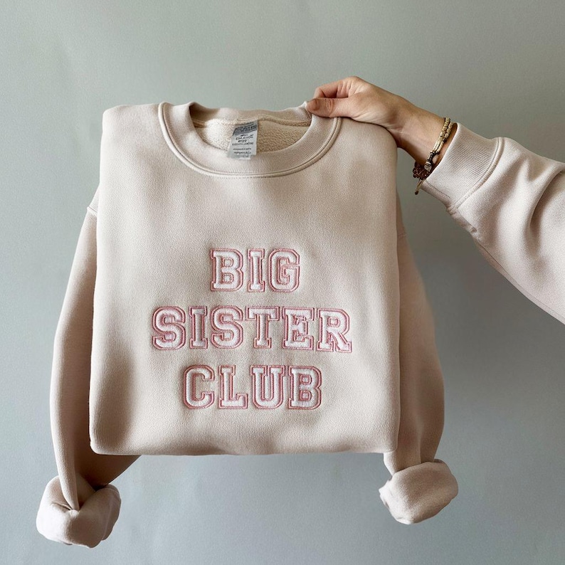 K&ouml;nnte beinhalten: Ein hellbeiger Pullover mit dem gestickten Text "BIG SISTER CLUB" in Rosa.