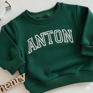 Puede incluir: Una sudadera verde oscuro con el nombre "ANTON" bordado en letras blancas. Tiene cuello redondo y pu&ntilde;os y dobladillo acanalados. La prenda est&aacute; hecha de un tejido suave.