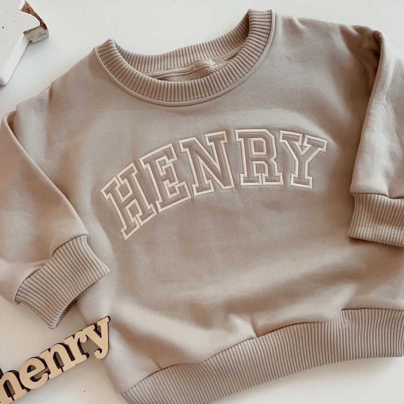 Puede incluir: Una sudadera beige de cuello redondo con el nombre "HENRY" bordado en letras blancas. La sudadera tiene pu&ntilde;os y dobladillo de canal&eacute;. La palabra "henry" tambi&eacute;n est&aacute; escrita con letras de madera en la parte inferior de la imagen.