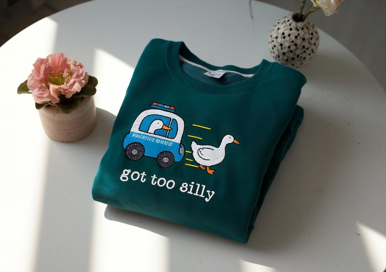 May include: Teal sweatshirt with a cartoon duck design. A blue police car with a duck inside is chasing another duck. The text "got too silly" is below the graphic. The sweatshirt is folded on a white surface.