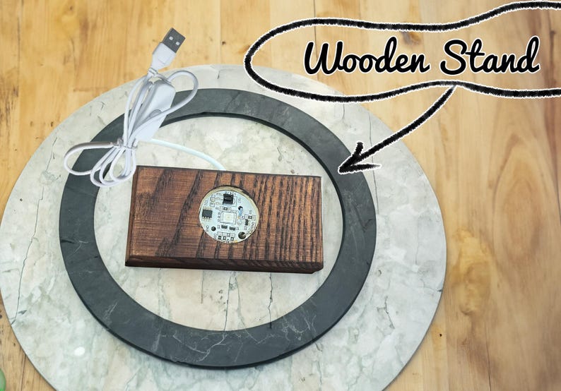 Puede incluir: Una peque&ntilde;a luz LED redonda blanca con una placa de circuito est&aacute; montada en un soporte de madera marr&oacute;n oscuro. El soporte est&aacute; sobre una superficie redonda blanca y negra. La luz est&aacute; conectada a un cable USB blanco.