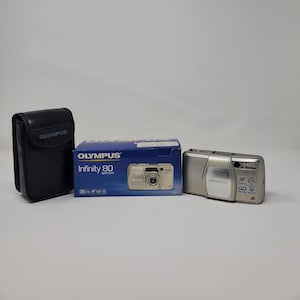 May include: An Olympus Infinity 80 camera with its original box and a black carrying case. The camera is silver and has the text "Olympus" on the front. The box is blue and has the camera's model name printed on it.