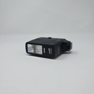 May include: A black camera flash unit with the text "SUPPOR" and "AUTO 211 THYRISTOR" printed on the side. The flash has a rectangular light source and a mounting bracket. The unit is on a white surface.