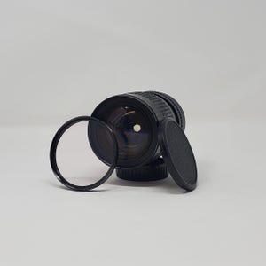 May include: A black camera lens with a clear filter and lens cap. The lens has a ribbed focus ring and a clear glass element. The filter is a black ring with clear glass. The lens cap is black and textured.