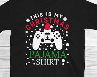 Könnte beinhalten: Schwarzes T-Shirt mit weißem Text, der "This is my Christmas Pajama Shirt" lautet, und einem weißen Videospiel-Controller mit einer roten Weihnachtsmütze oben drauf. Der Controller ist von weißen Schneeflocken umgeben.