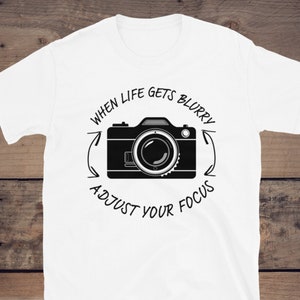 May include: White t-shirt with a black camera graphic and the text "When life gets blurry adjust your focus".
