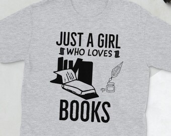 Può includere: Maglietta grigia con testo nero che recita "Just a girl who loves books". Il design include una pila di libri, una penna stilografica e un calamaio.