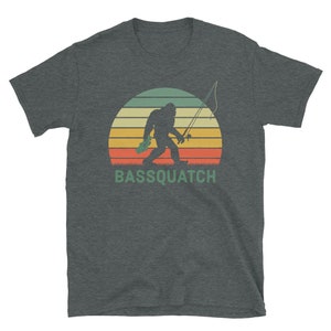 May include: A dark grey t-shirt featuring a retro sunset graphic with Bigfoot holding a fishing rod and a fish. The text "BASSSQUATCH" is printed below the graphic.