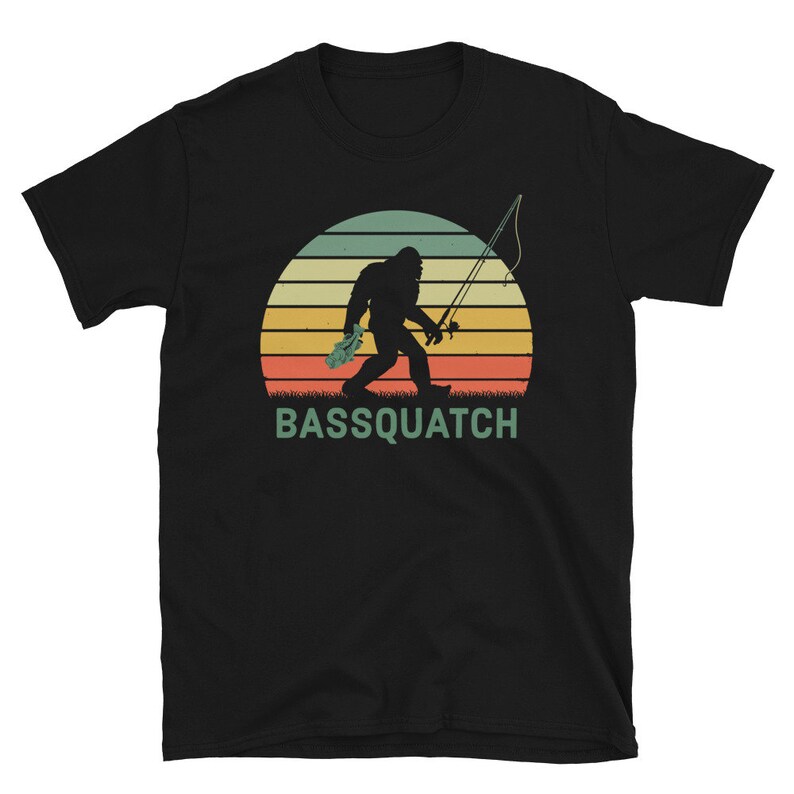 May include: A black t-shirt featuring a retro-style illustration of Bigfoot holding a fishing rod and a fish. The illustration is set against a sunset background with the text "BASSSQUATCH" below.