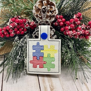May include: White leather keychain with a blue, red, yellow, and green puzzle piece design. The keychain has a silver metal ring.