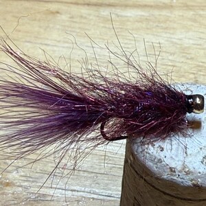 May include: A close-up of a handmade fishing fly. The fly has a dark purple tail and body, a black hook, and a gold-coloured bead head. The fly is designed to mimic an insect and is used for fly fishing.