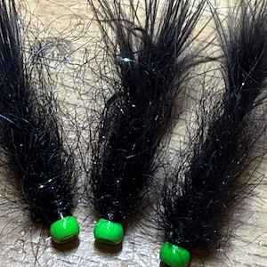 May include: Three black fishing flies with green bead heads. The flies have a fluffy, textured appearance with strands of black material and a touch of sparkle. The green beads are a bright, solid color.