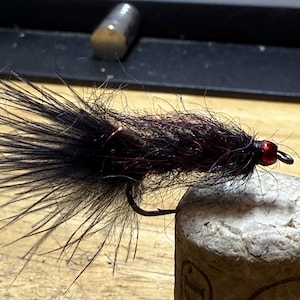 May include: A handmade fly fishing lure with a black body and red accents. The lure features a curved hook and a red bead head. It is resting on a cork surface, showcasing the craftsmanship and detail of the fly.