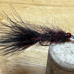 May include: A handmade fishing fly with a black body and red accents. The fly has a red bead head and is tied with fine, dark fibers. It is resting on a cork surface, showcasing its intricate design and craftsmanship.