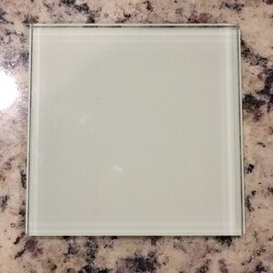 Puede incluir: Un posavasos cuadrado de vidrio esmerilado con una superficie lisa y blanca. El posavasos tiene un borde delgado de plata.