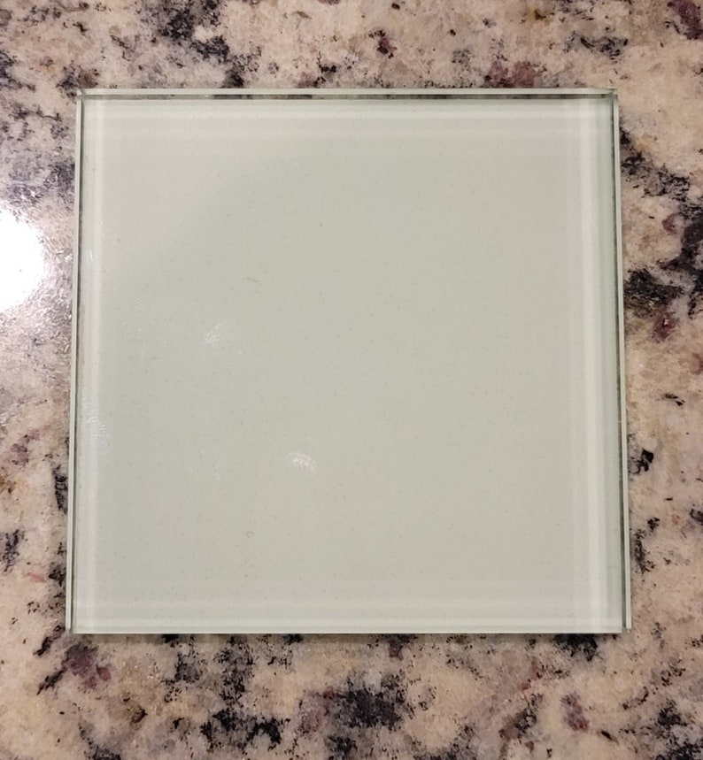 Puede incluir: Un posavasos cuadrado de vidrio esmerilado con una superficie lisa y blanca. El posavasos tiene un borde delgado de plata.