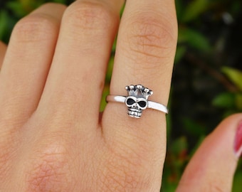 Puede incluir: Un anillo de plata con un pequeño diseño de calavera. La calavera lleva una corona.