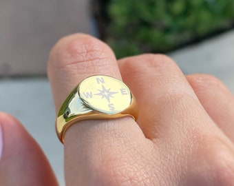 Puede incluir: Un anillo de sello de oro con un diseño de brújula. La brújula presenta una estrella blanca y las letras N, E, S y W.