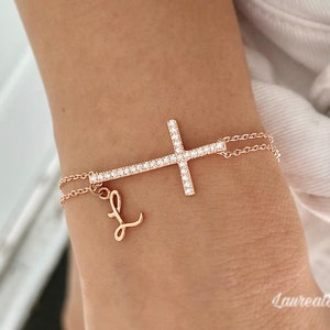 Puede incluir: Una pulsera de oro rosa con un delicado dije de cruz y un dije de letra 'L' en cursiva. El dije de cruz está incrustado con pequeñas piedras transparentes.