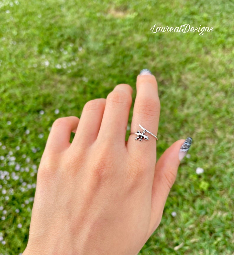 May include: A silver ring with a small lizard design. The ring is wrapped around the finger and has a thin band.