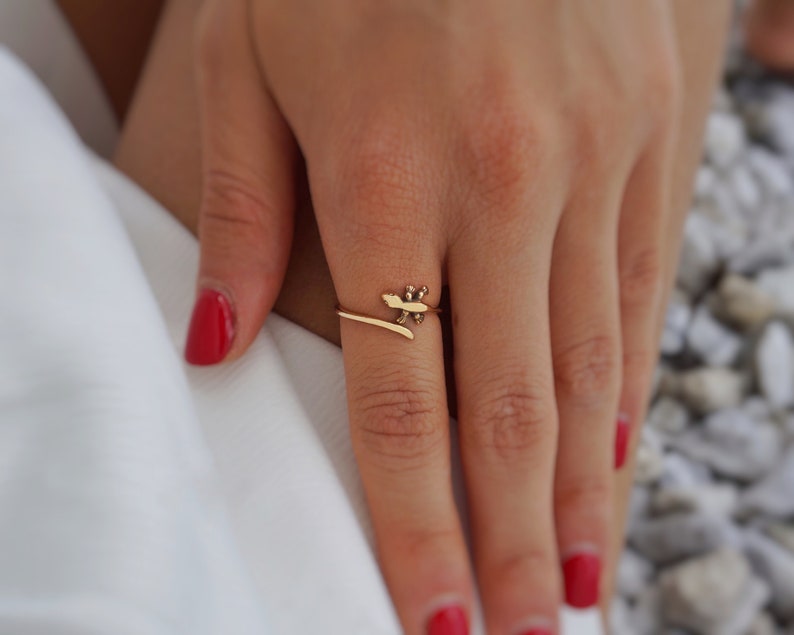 May include: A gold ring with a delicate design of a stylized leaf. The ring is worn on a finger with red nail polish.