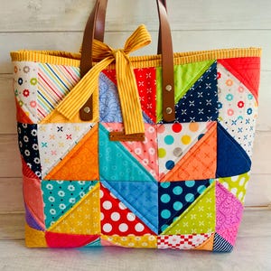 May include: A patchwork tote bag in a vibrant mix of colours and patterns, including red, blue, yellow, and green. It features brown leather handles and a yellow striped bow. The bag is approximately 35 cm wide and 30 cm tall.