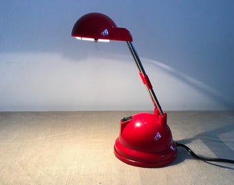 Könnte beinhalten: Eine leuchtend rote Schreibtischlampe mit kuppelförmigem Schirm, einem verstellbaren Hals in Chromoptik und einem abgerundeten Sockel. Die Lampe verfügt über einen schwarzen Schalter auf der Oberseite des Sockels und ein schwarzes Stromkabel.